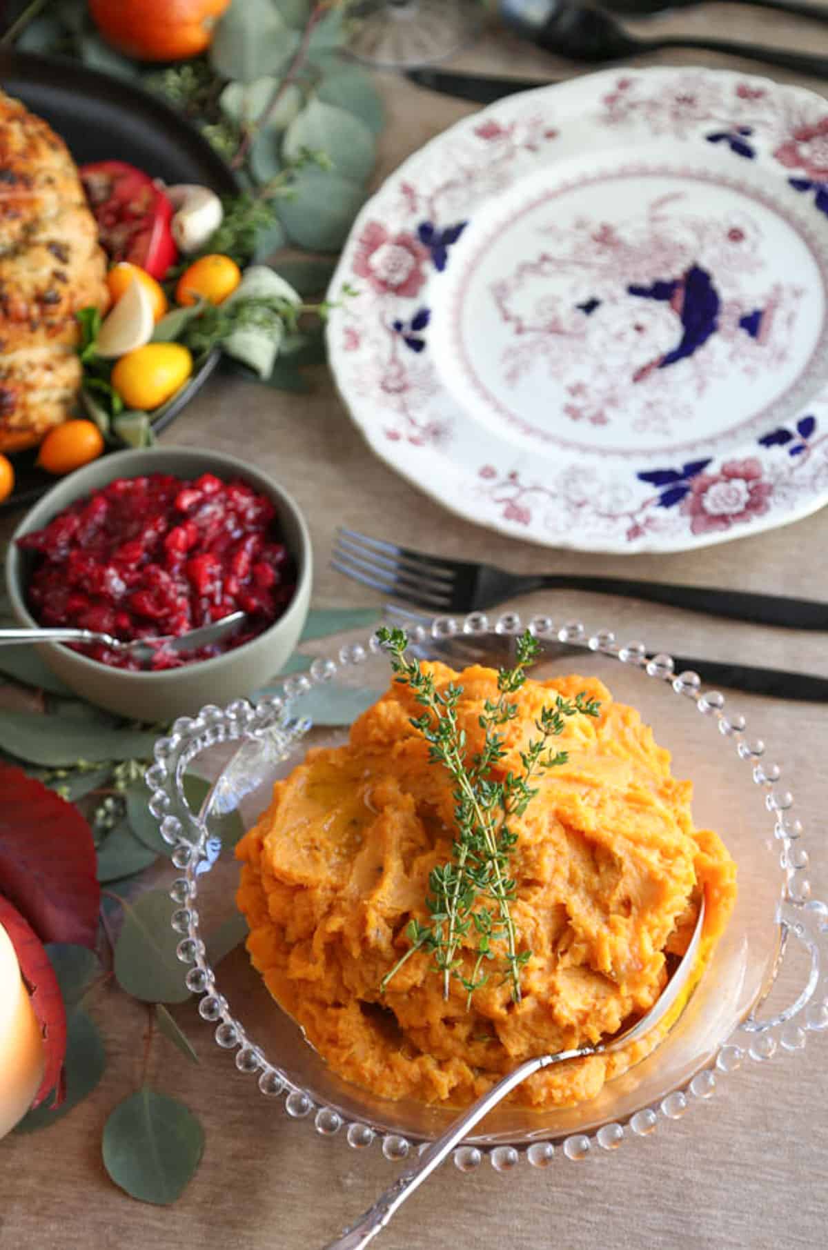 mashed sweet potatoes topped with fresh thyme on a table set for dinner with china plates, turkey, and cranberry sauce.