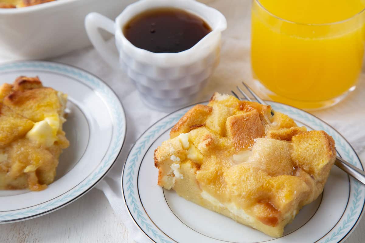 slices of french toast casserole on plates next to a mini pitcher of maple syrup and a glass of orange juice.