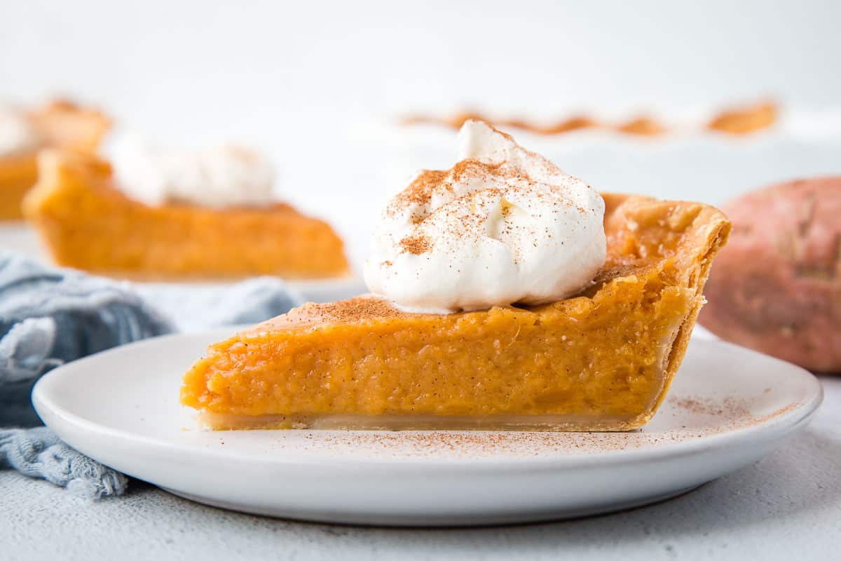 slice of sweet potato pie on a white plate, topped with whipped cream, with another slice in the background.