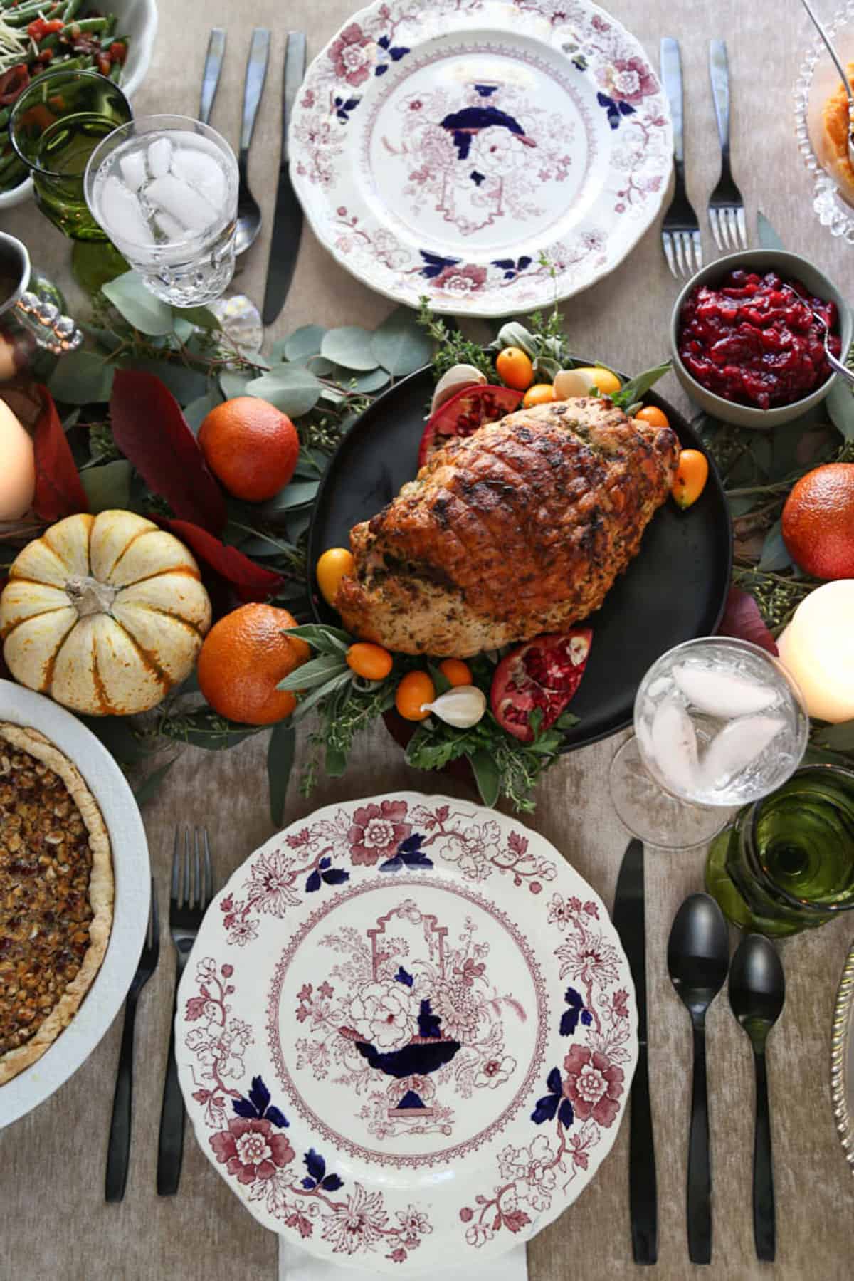 table set with china plates, mini pumpkins, blood oranges, and leaves, with a turkey breast on a black platter in the center.