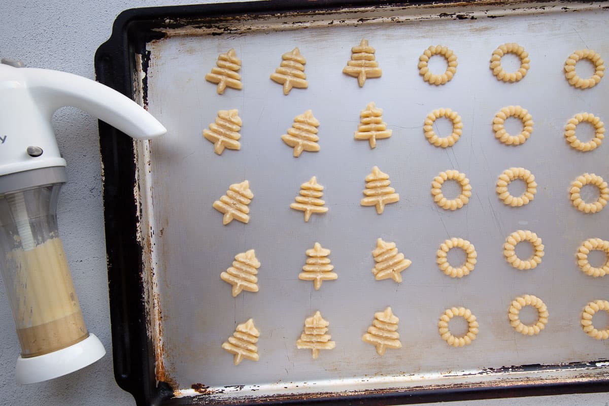 a cookie press next to a baking sheet of piped spritz cookies that are uncooked.