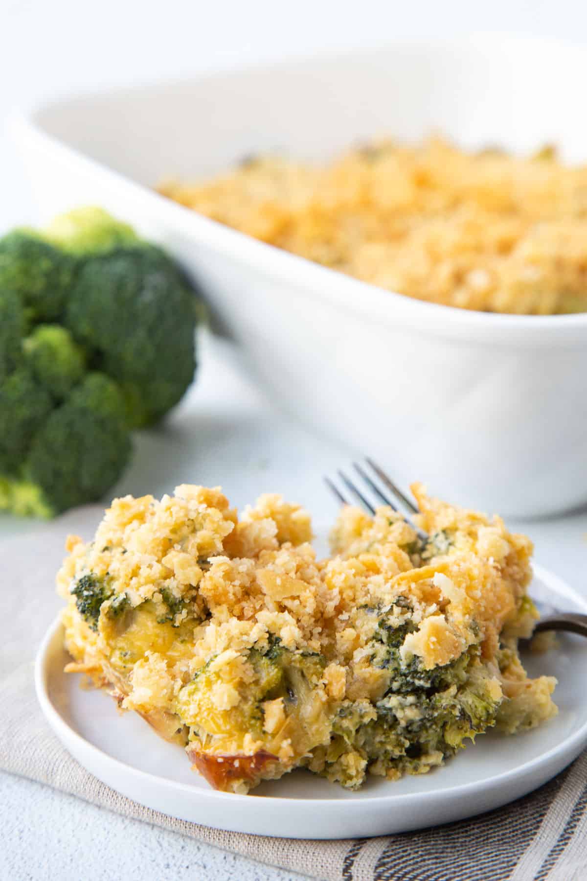 slice of velveeta broccoli casserole on a white plate in front of a white casserole dish with more casserole.
