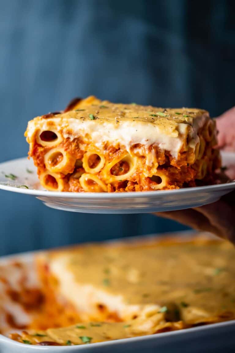 hand holding a plate of baked ziti near a casserole dish full of more ziti.