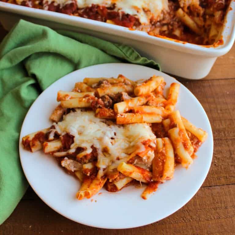 baked ziti on a white plate, next to a casserole dish and a green cloth napkin.