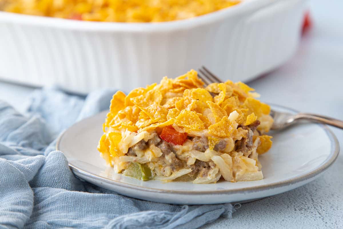 slice of hash brown casserole on a white flat with a fork, topped with corn flakes.