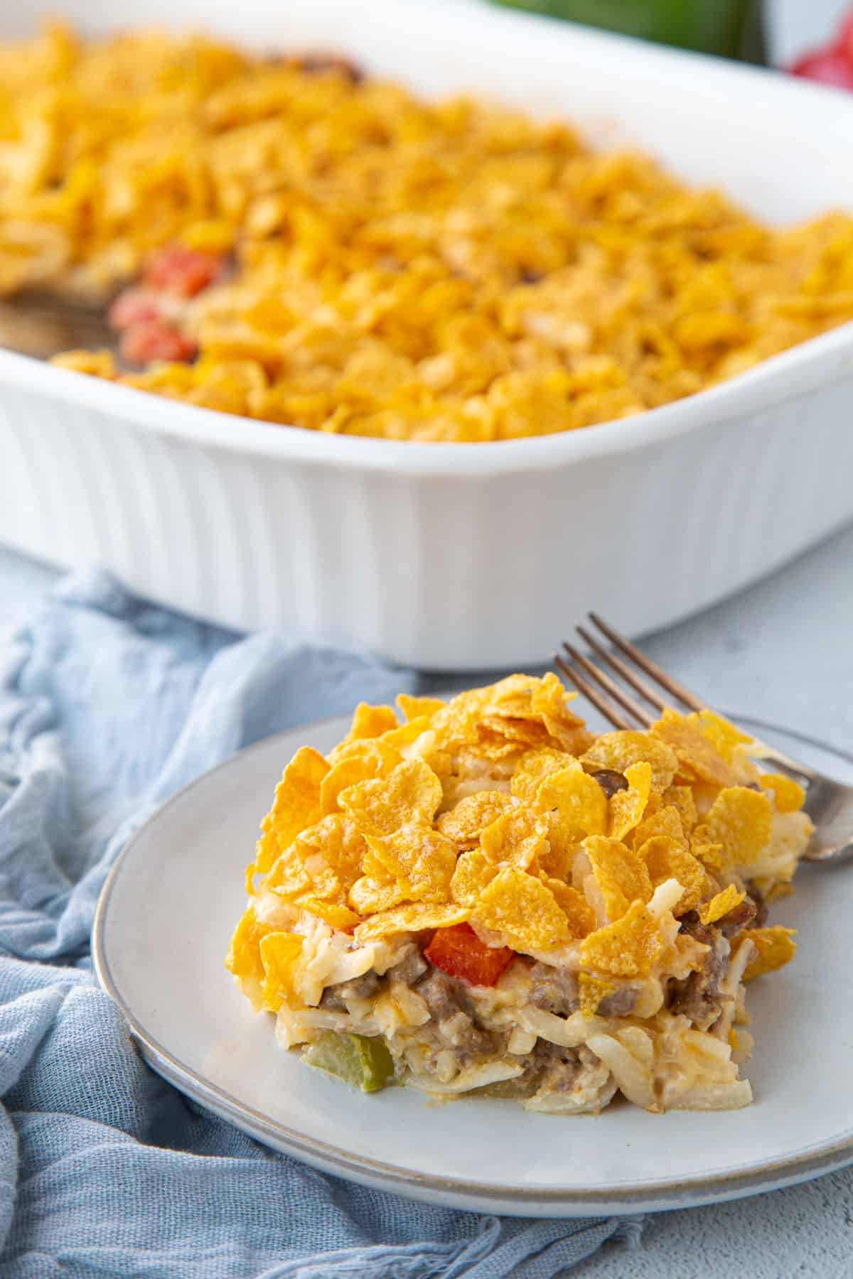 slice of hash brown casserole on a white plate in front of a white casserole dish filled with more hash brown casserole.