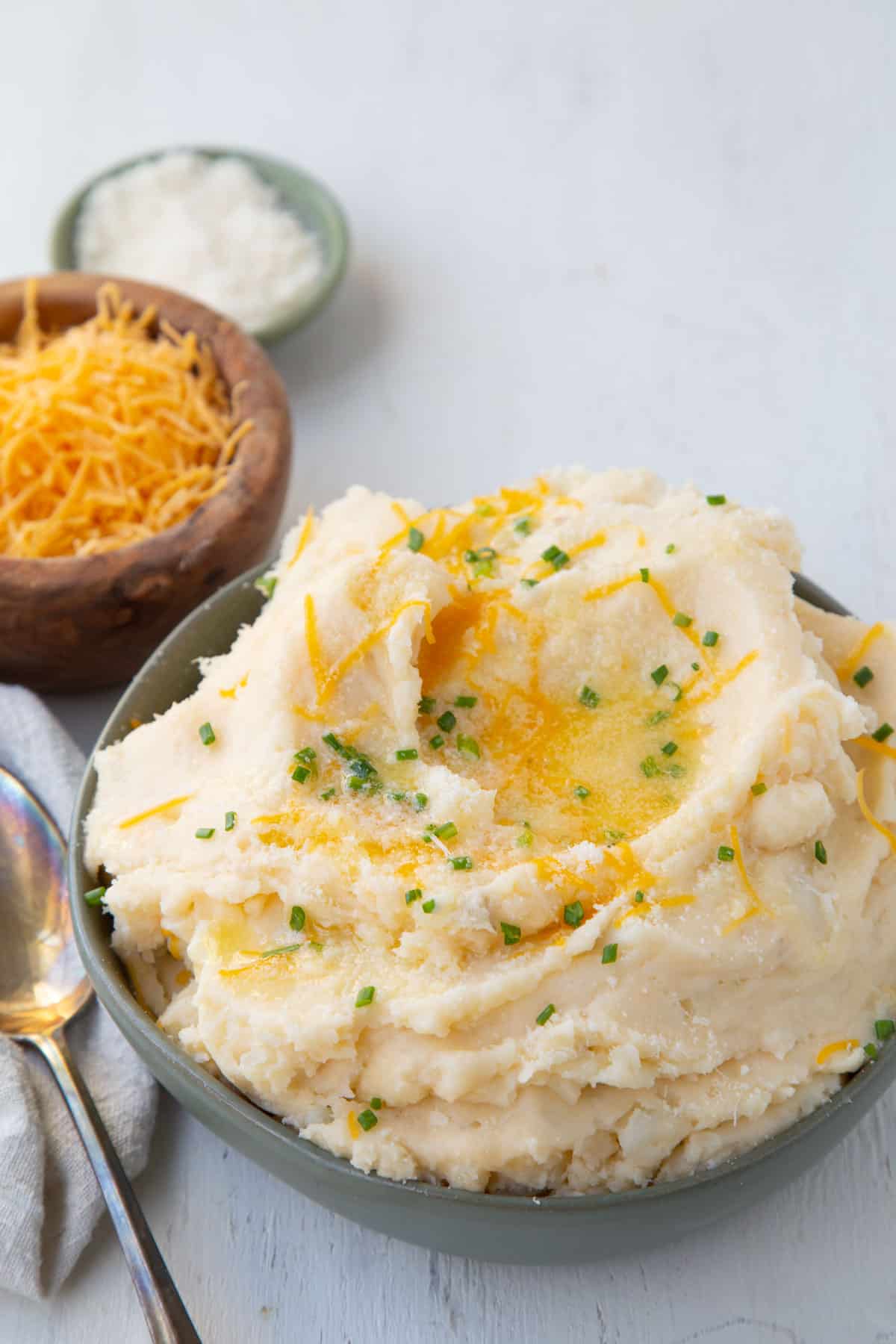 cheesy mashed potatoes topped with chives and butter in a green bowl, next to smaller bowls of cheddar and parmesan.