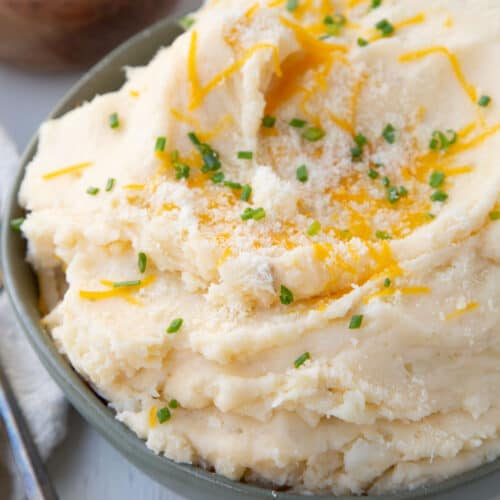 cheesy mashed potatoes in a green bowl, topped with chives and shredded cheddar.