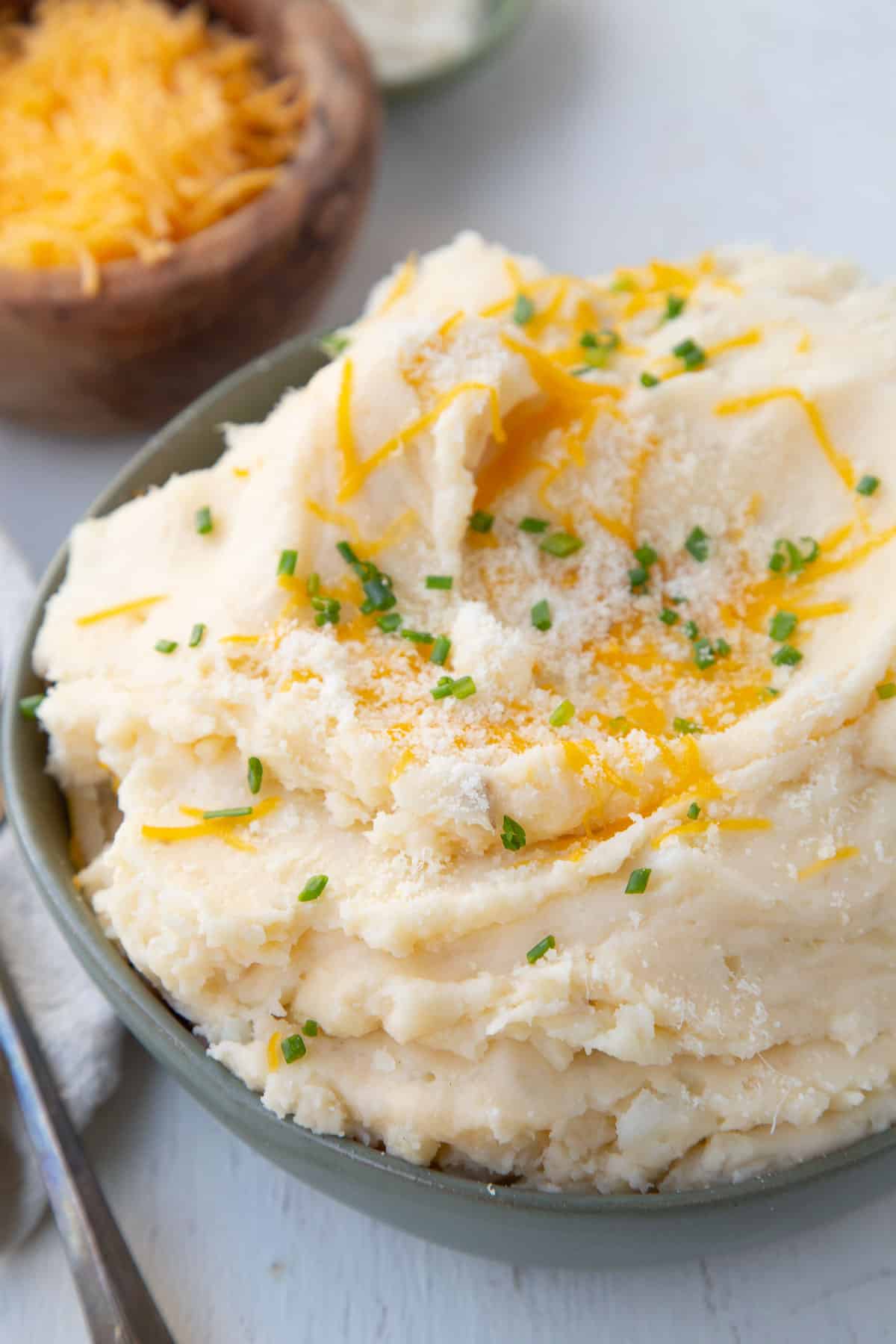 cheesy mashed potatoes topped with chives in a green bowl, next to a small wooden bowl of shredded cheddar.