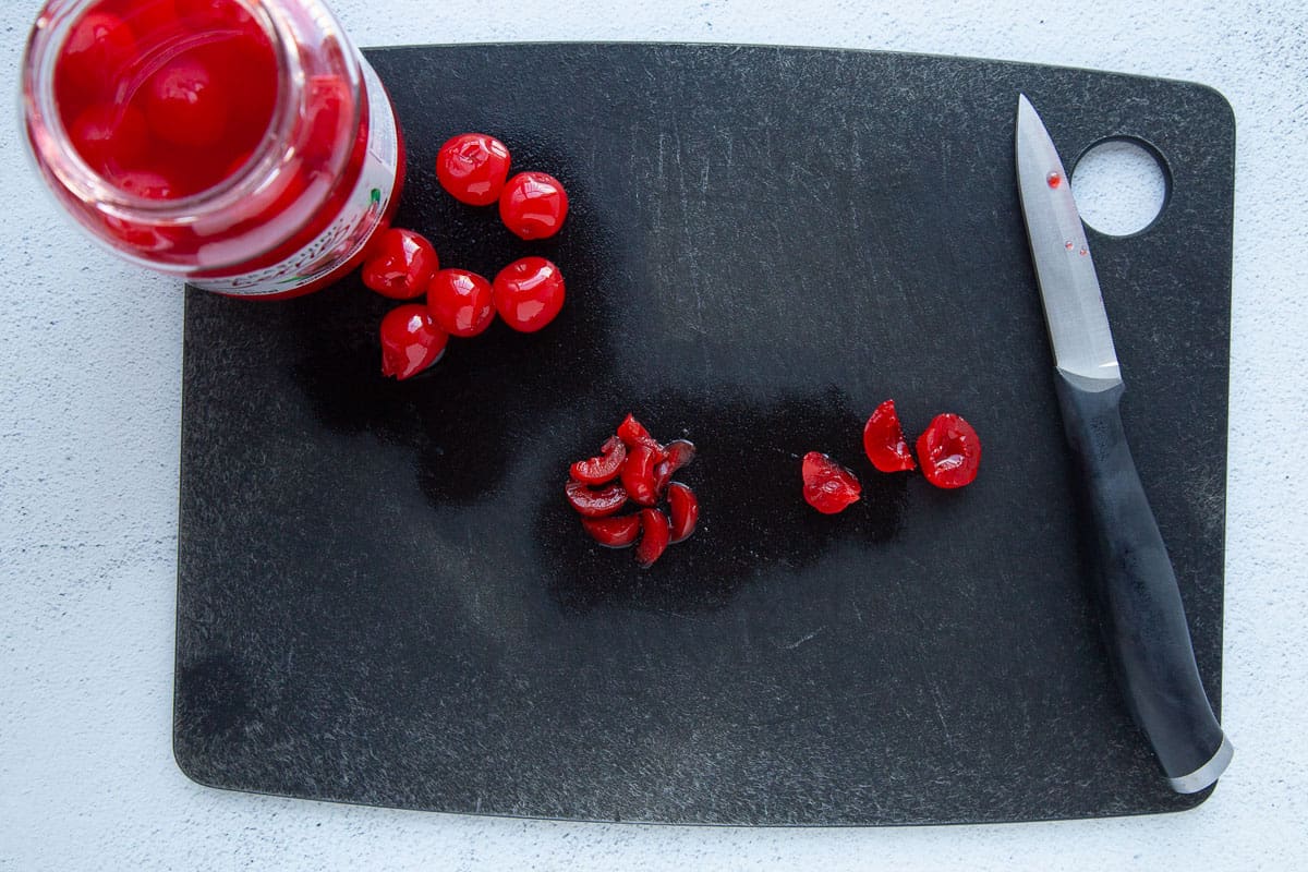 sliced and whole maraschino cherries on a black cutting board with a paring knife.