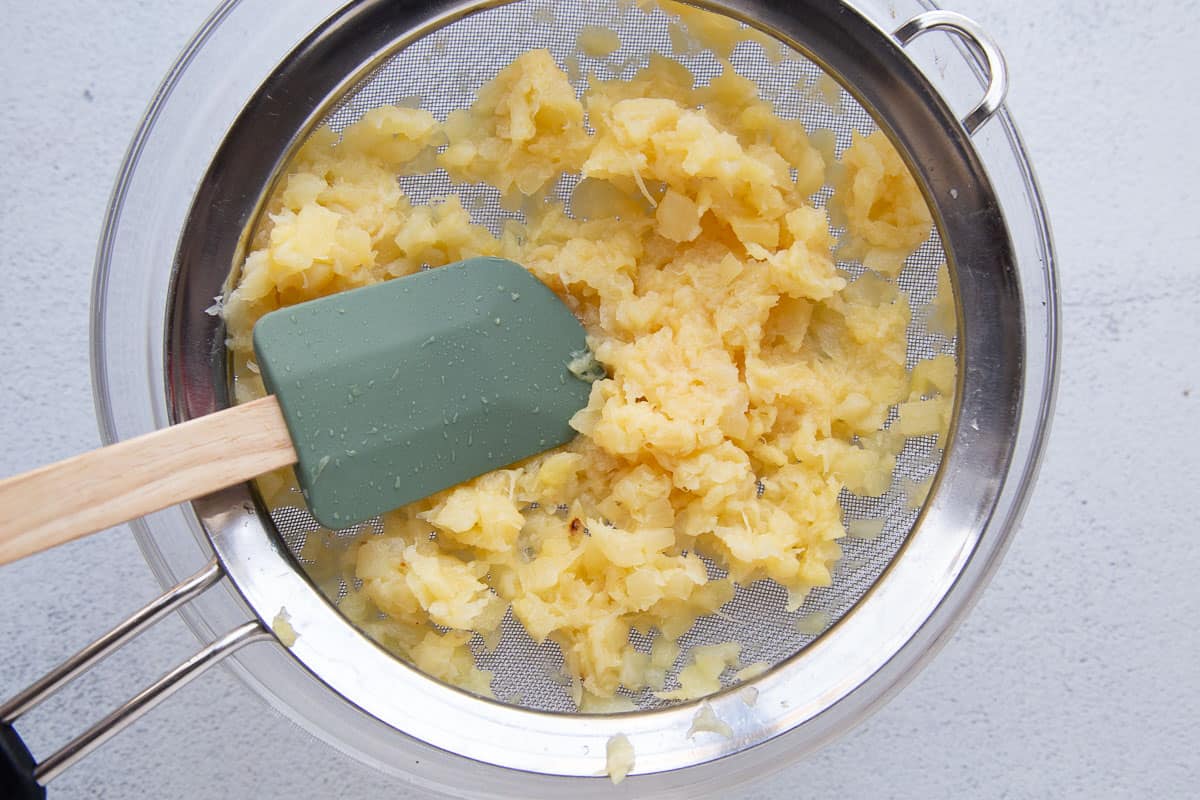 spatula pressing on crushed pineapple in a mesh colander.