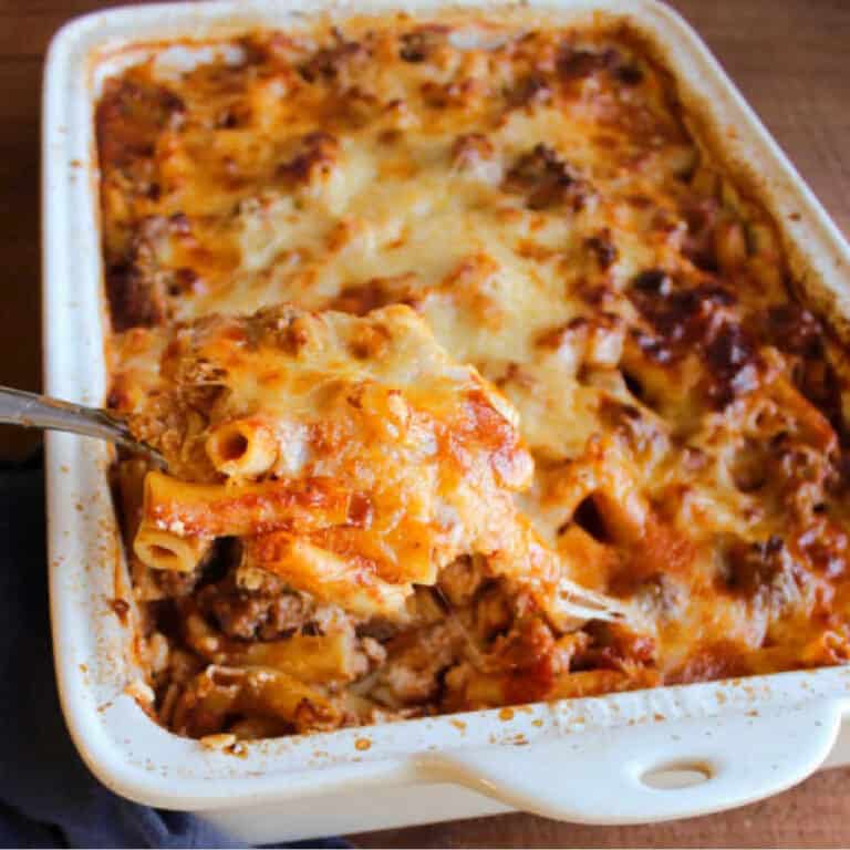 spoon lifting a spoonful of cheesy baked ziti from a white casserole dish.