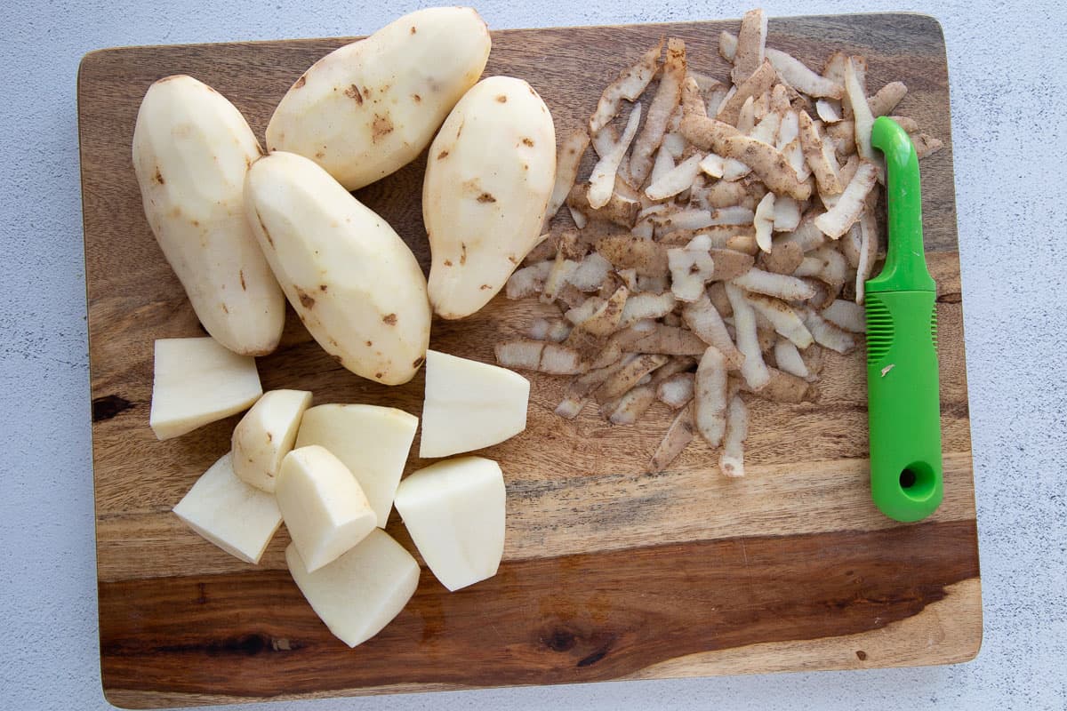 peeled whole potatoes and peeled chopped potatoes on a cutting board with potato peels and a peeler.