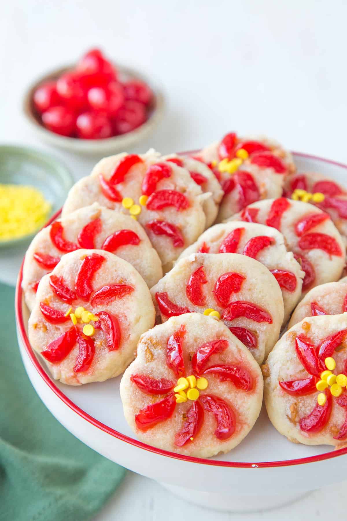 white cake stand with a red rim holding cookies topped with maraschino cherries to resemble poinsettias. 