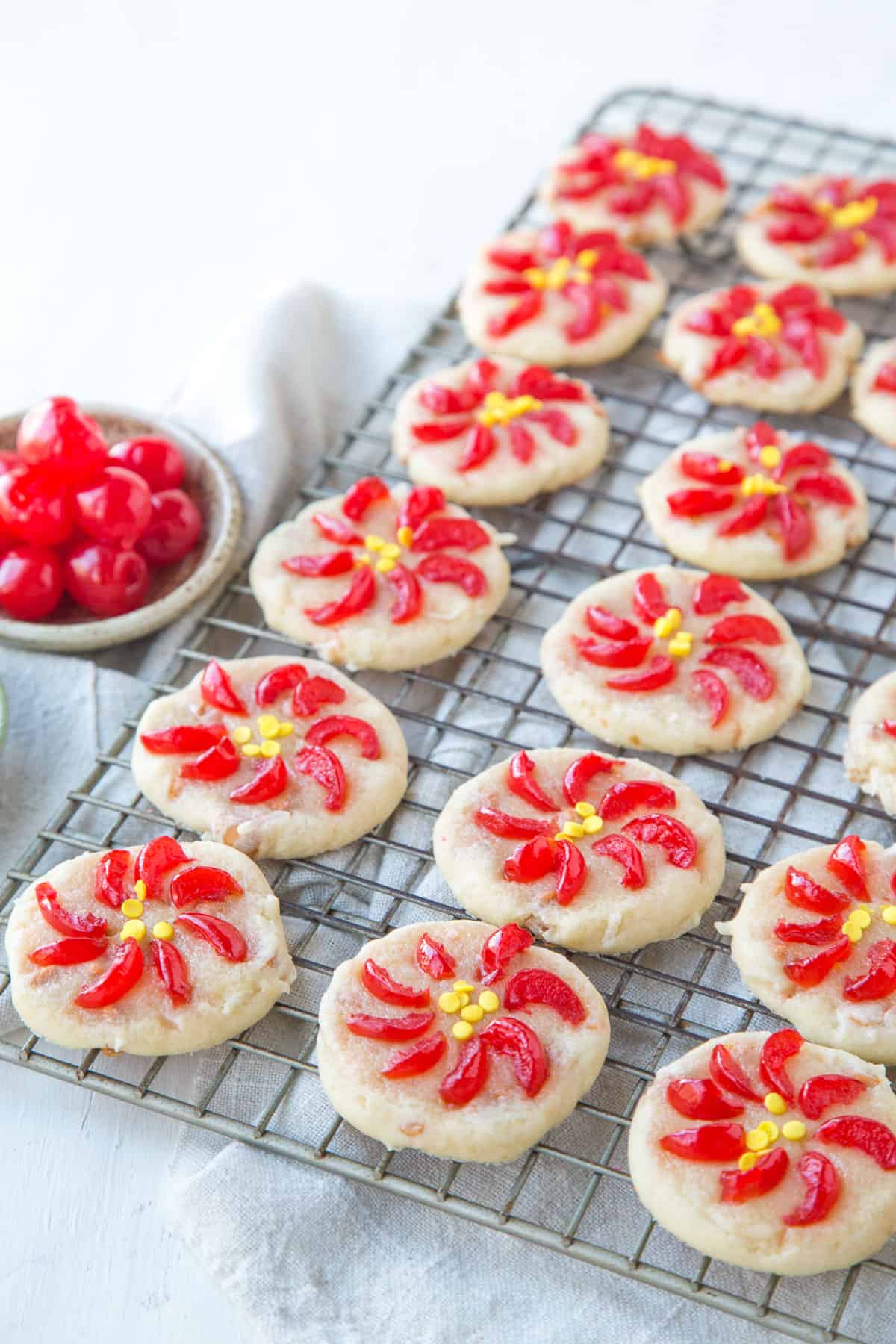 round beige cookies decorated with sliced maraschino cherries and sprinkles to resemble poinsettias, on a wire rack.
