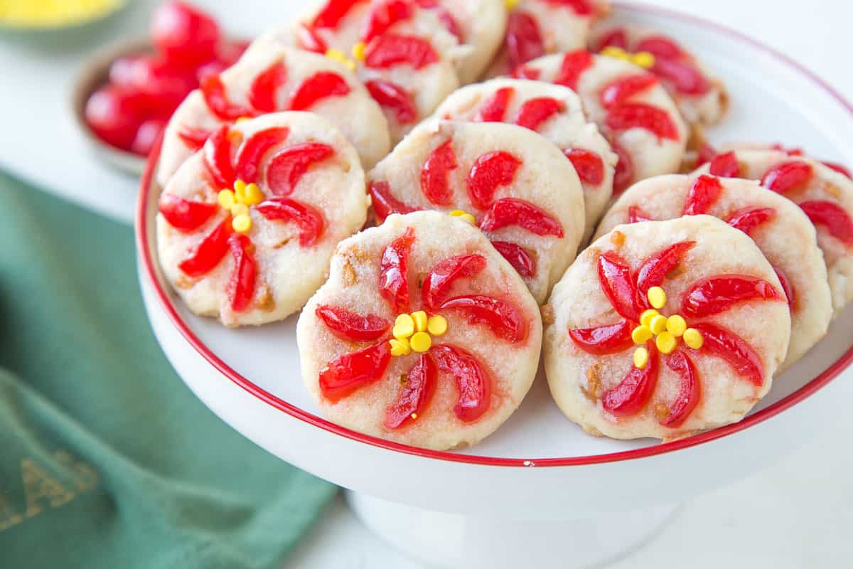 cookies with maraschino cherries sliced and arranged to look like poinsettias on a white cake stand.