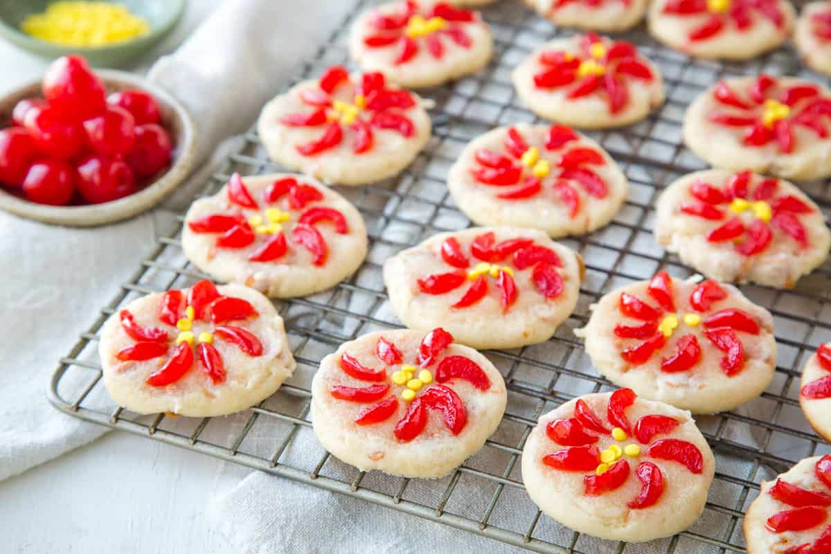 poinsettia cookies on a wire rack next to a small bowl of maraschino cherries.
