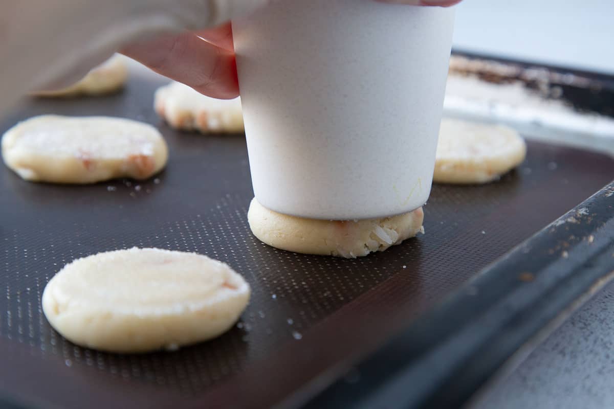 hand pressing a glass onto a piece of raw cookie dough on a baking sheet.