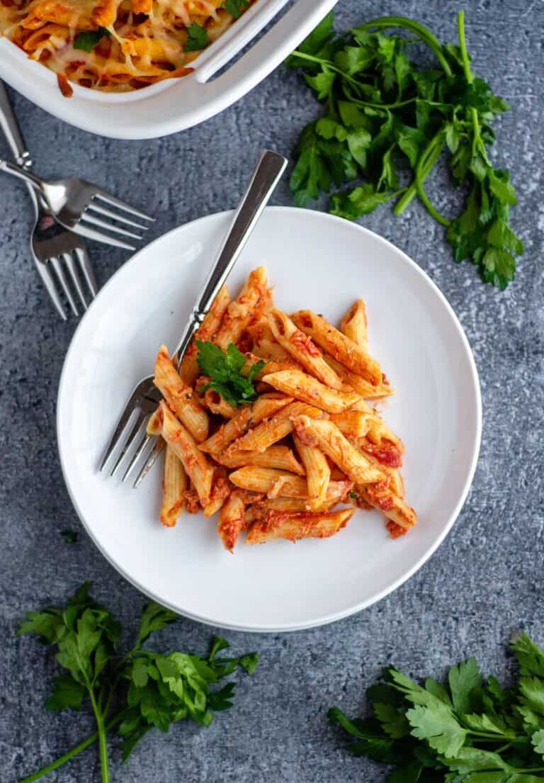 baked ziti on a white plate next to a whole casserole dish and bunches of parsley.