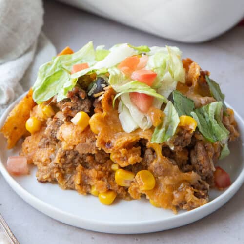 taco casserole with fritos on a small white plate, topped with lettuce and tomato.