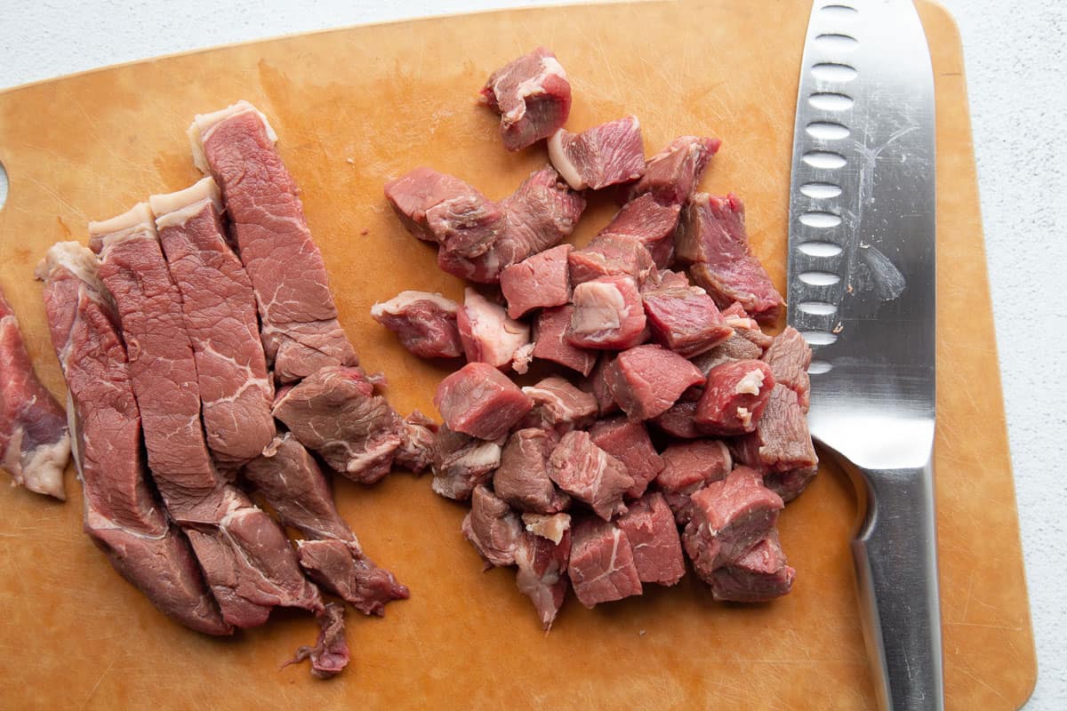 cubes and strips of beef sirloin on a cutting board next to a chef's knife.