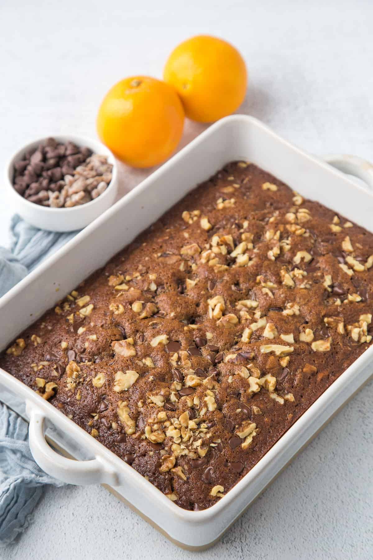 date cake in a white casserole dish next to a small bowl of dates and two oranges.