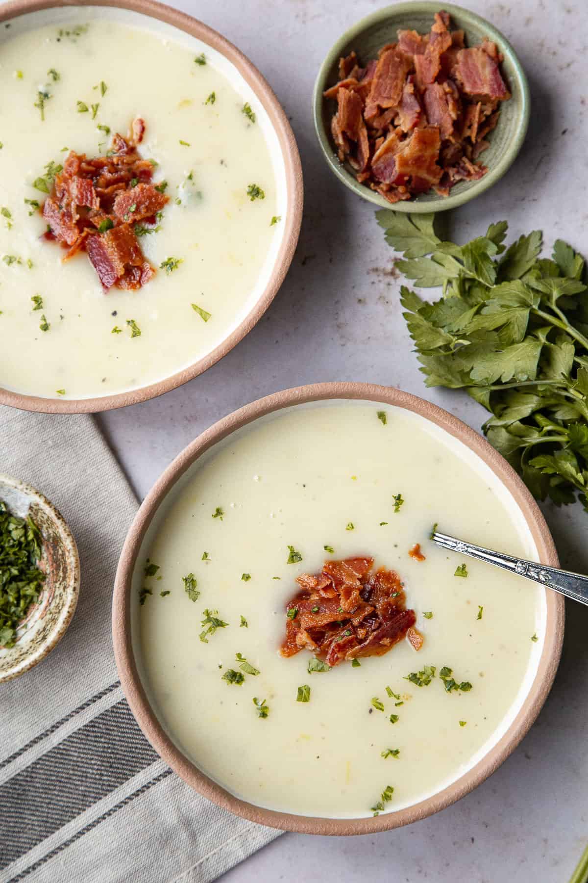 two bowls of potato soup topped with bacon and parsley, next to a small bowl of chopped bacon.