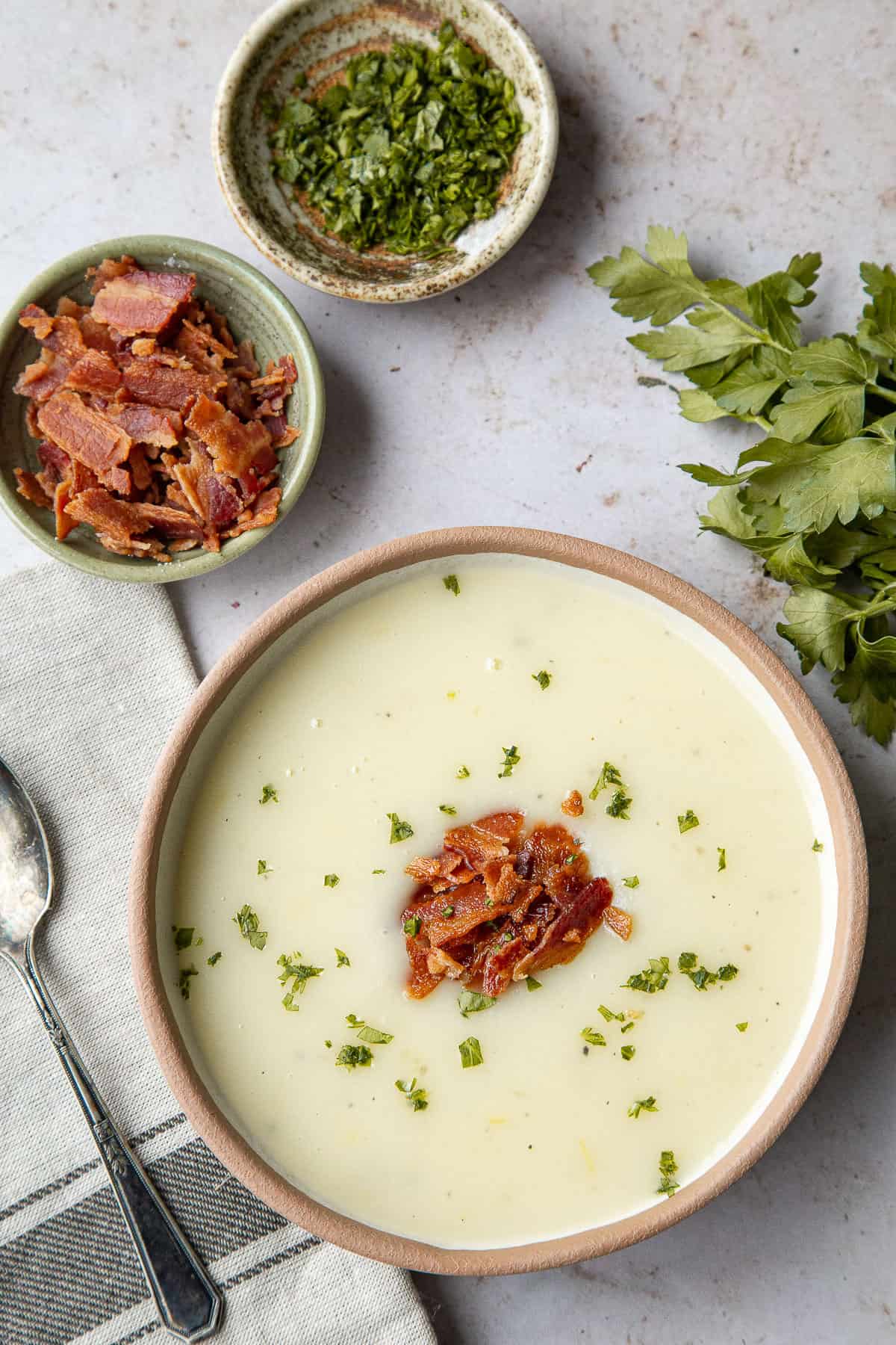 bowl of potato soup with two small bowls of bacon and chopped parsley next to it.