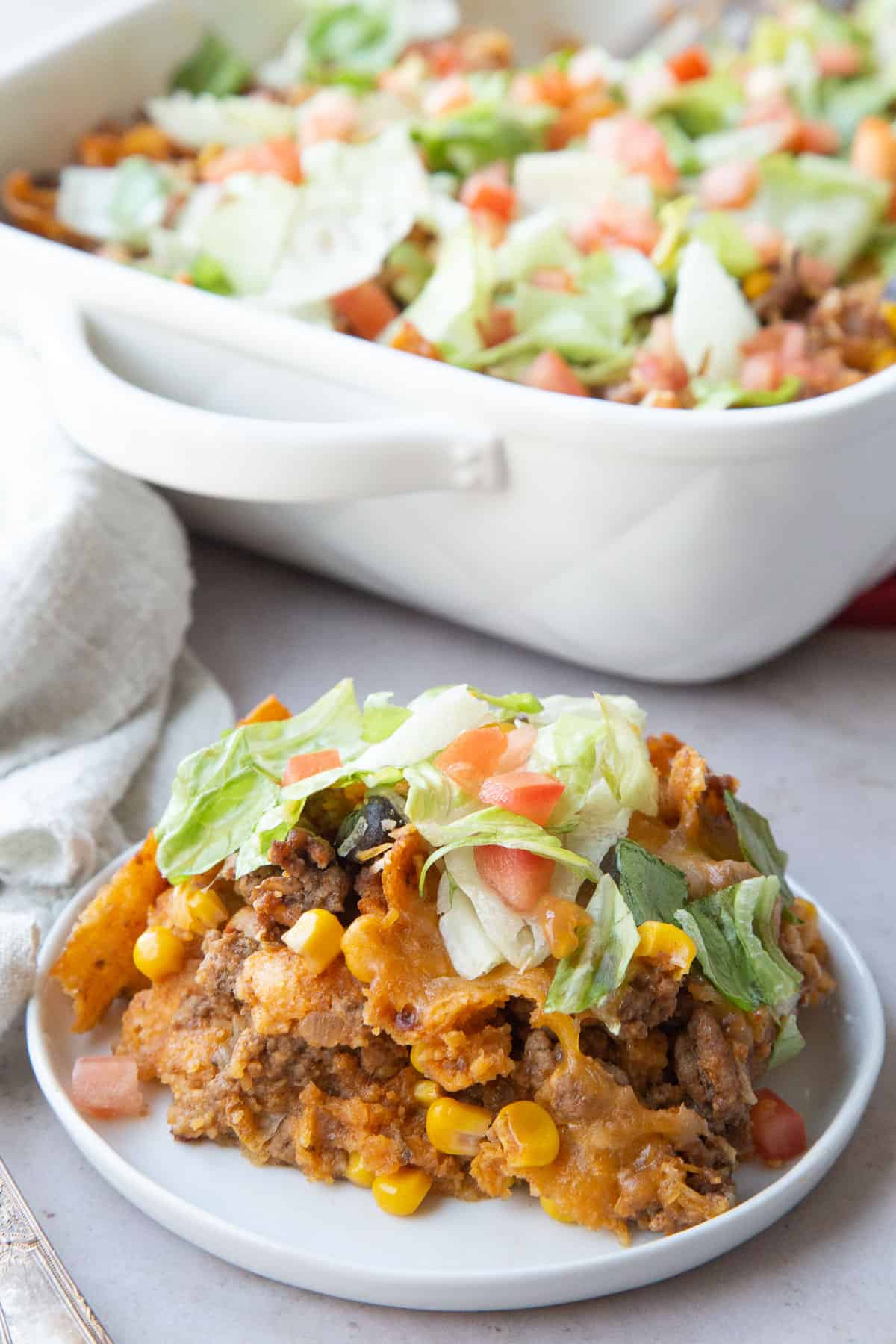 slice of taco casserole on a white plate, in front of a full casserole dish.