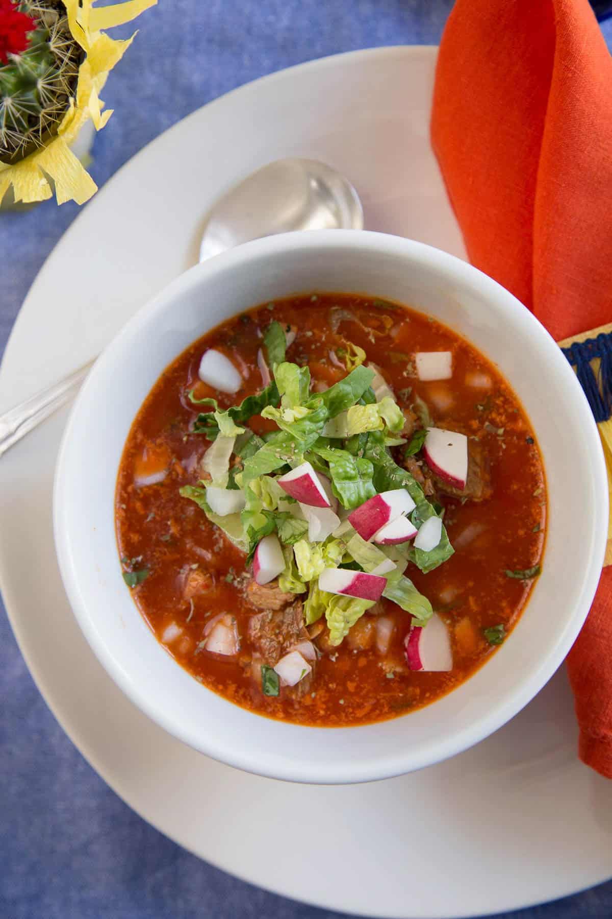 white bowl of red pozole on a white plate next to a bright orange napkin.