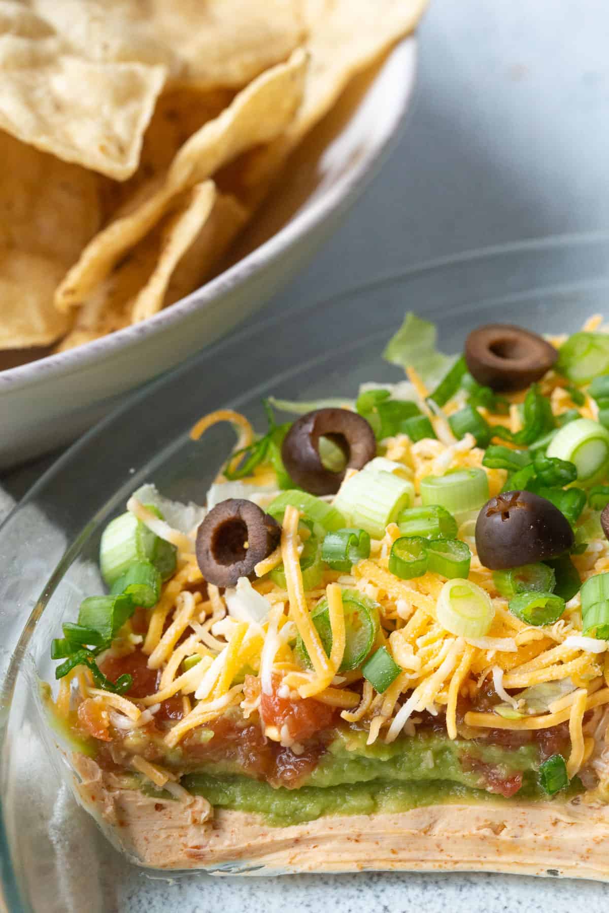 7 layer taco dip with cream cheese, guacamole, salsa, cheese, lettuce, green onions, and black olives in a glass pie dish next to a bowl of chips.