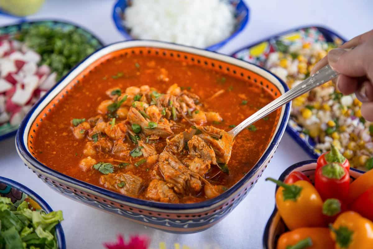 hand lifting a silver spoonful of red pozole from a decorative dish.