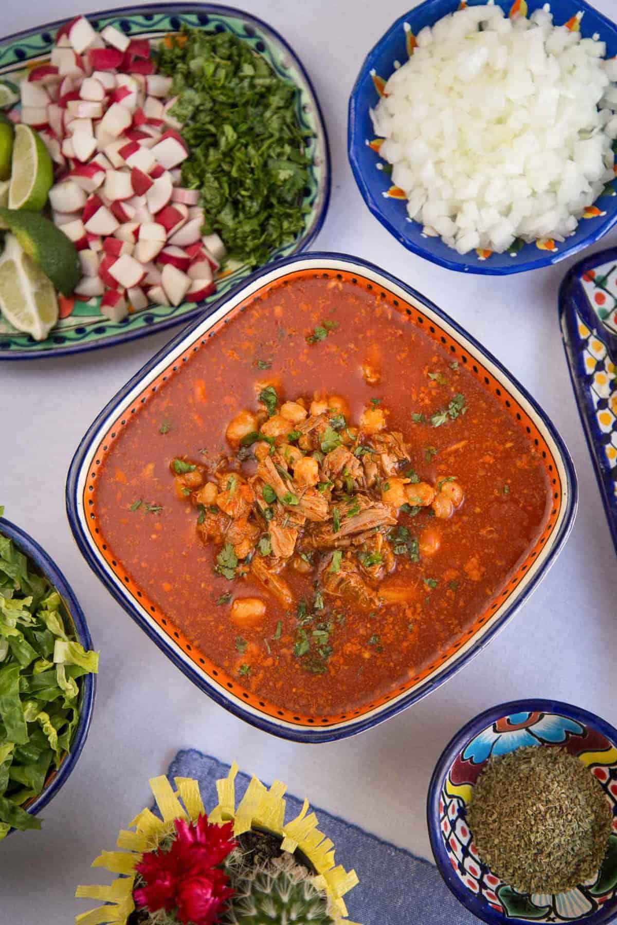 large bowl of red pozole surrounded by dishes of toppings including chopped white onion, radish, shredded lettuce, and lime slices.