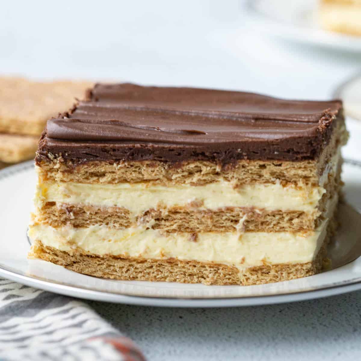 slice of layered chocolate eclair cake on a white plate, topped with chocolate frosting.