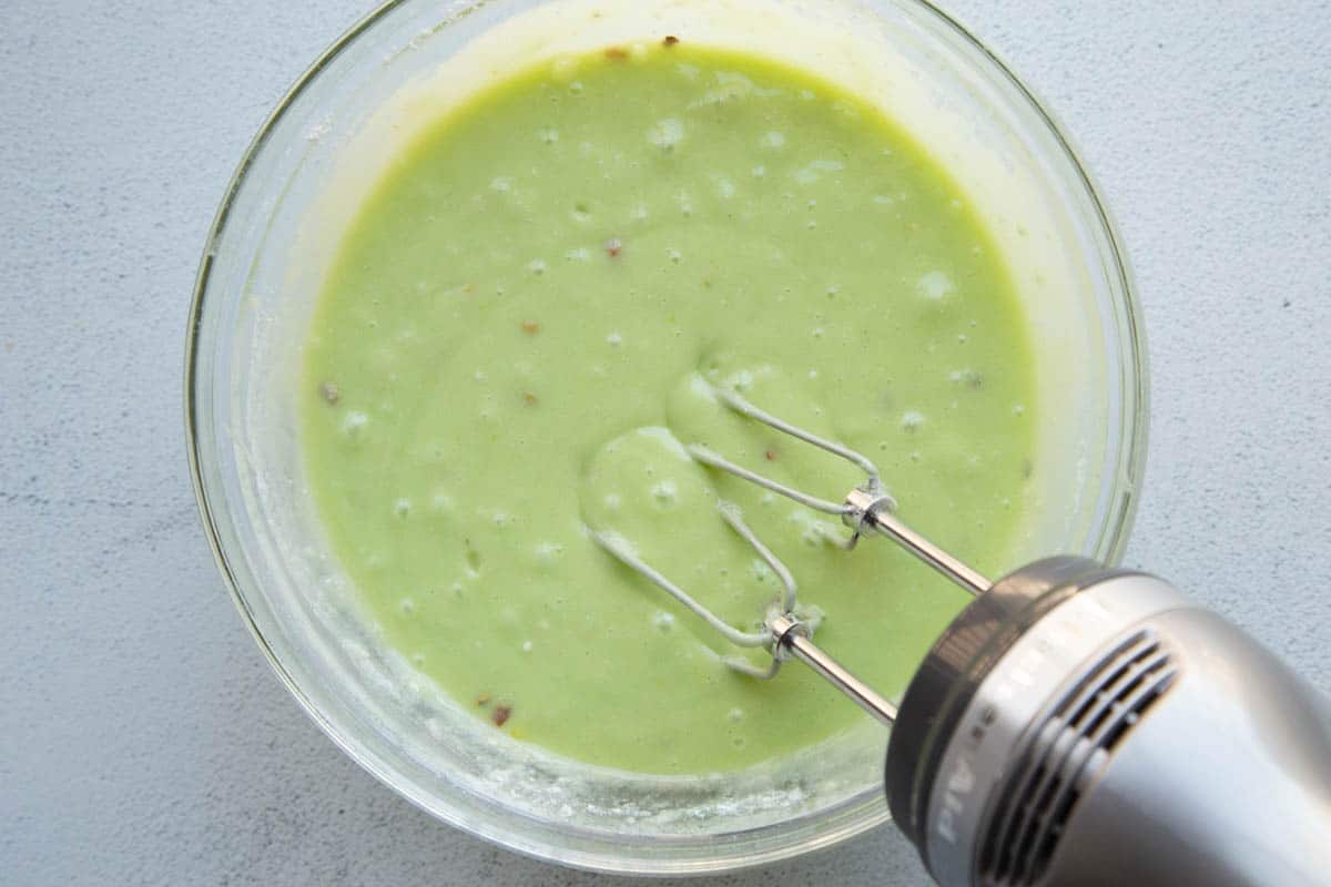 green cake batter in a glass bowl with a handheld mixer.