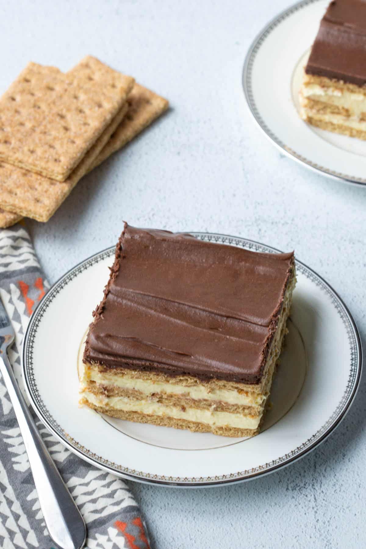 piece of chocolate eclair cake on a white plate, next to a printed napkin, a fork, and a stack of graham crackers.