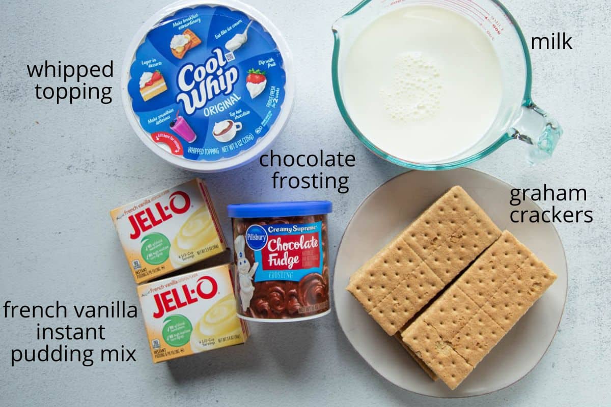 graham crackers, whipped topping, boxes of vanilla pudding, a can of chocolate frosting, and a glass cup of milk.