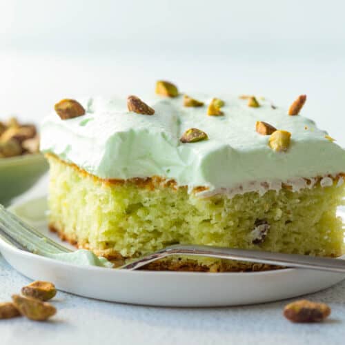 slice of pistachio cake topped with pistachio frosting and chopped pistachios on a white plate.