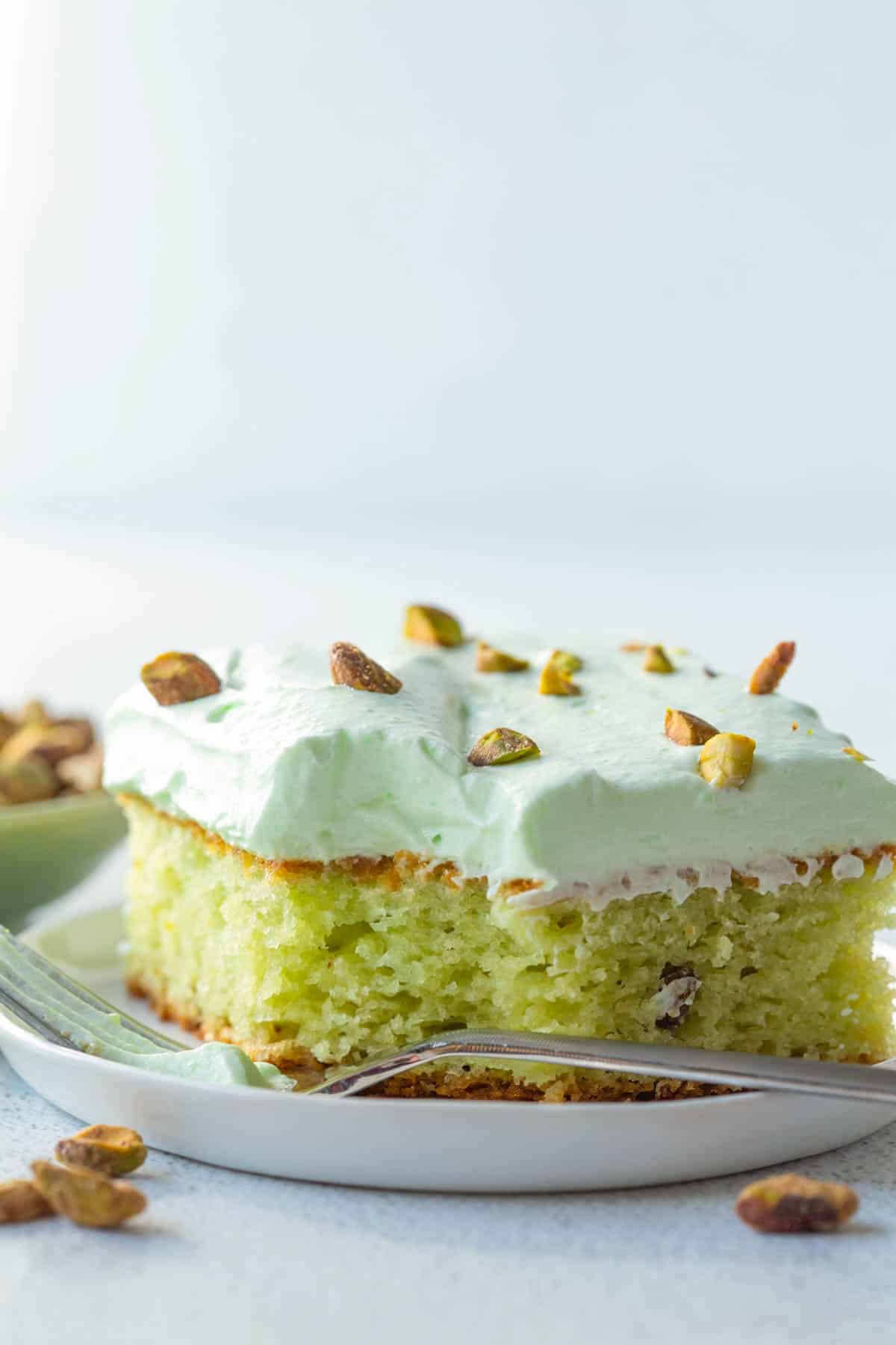 slice of pistachio cake with pudding mix on a white plate, topped with a pistachio frosting and chopped pistachios.