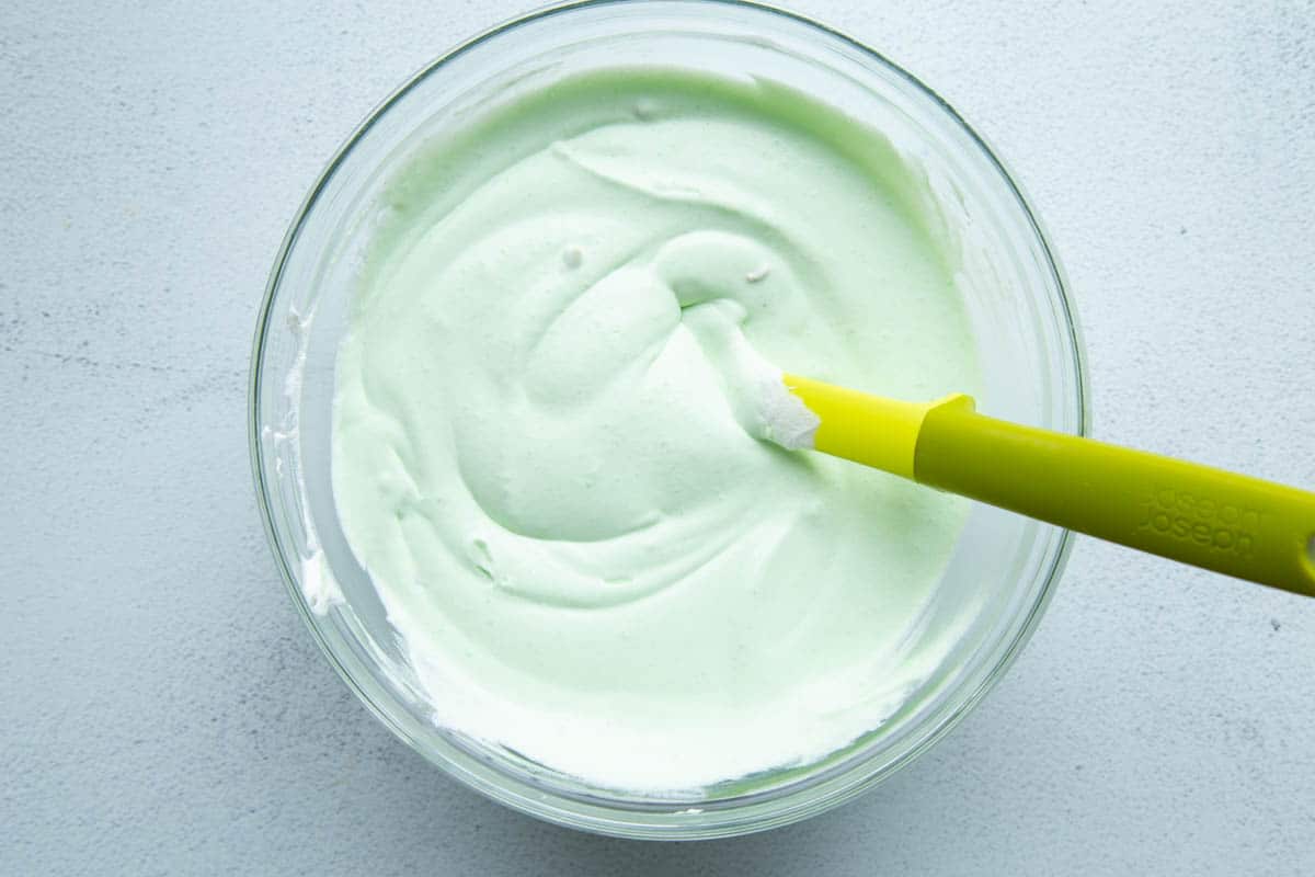 green fluffy cool whip frosting in a glass bowl with a yellow spatula.