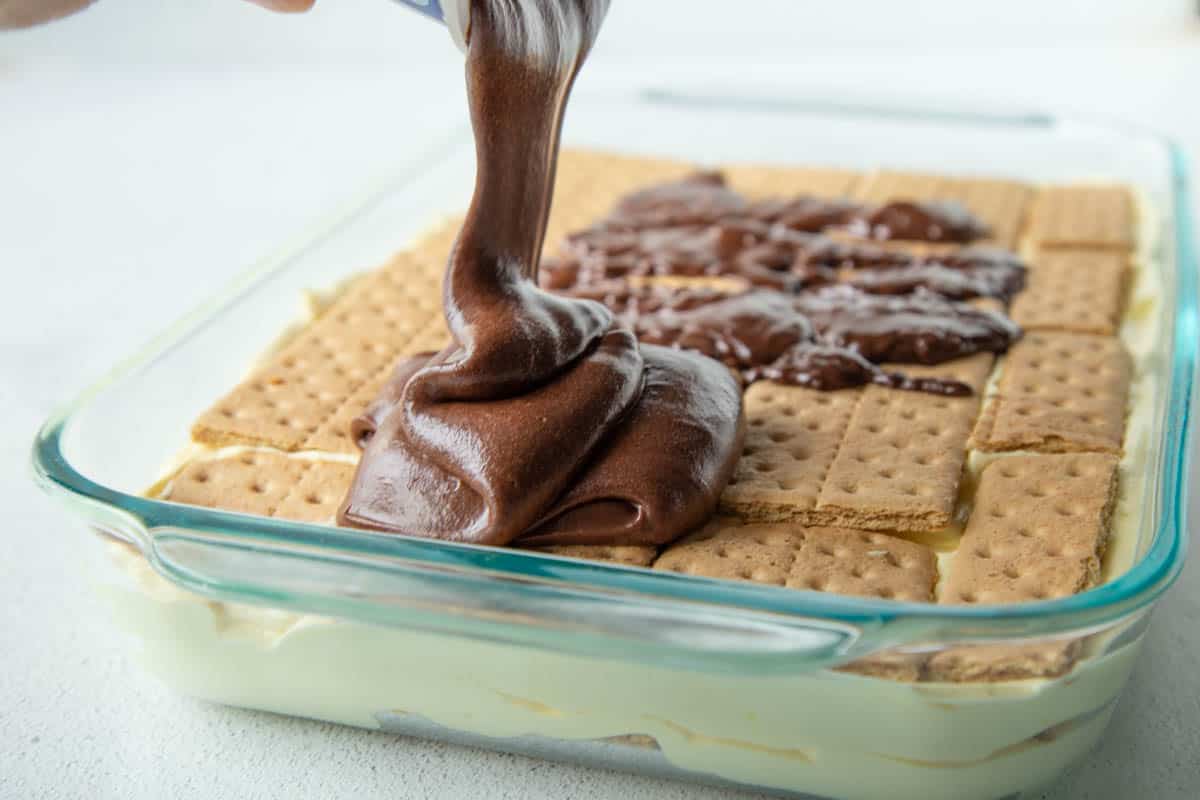 hand pouring chocolate frosting onto a layer of graham crackers.