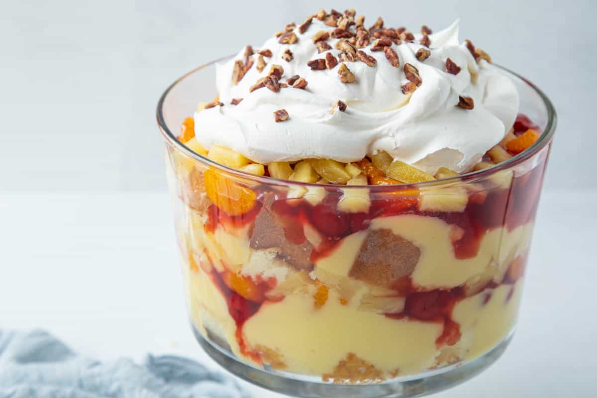 glass trifle dish filled with layers of cake cubes, vanilla pudding, cherry pie filling, and fruit, topped with whipped topping and pecans.