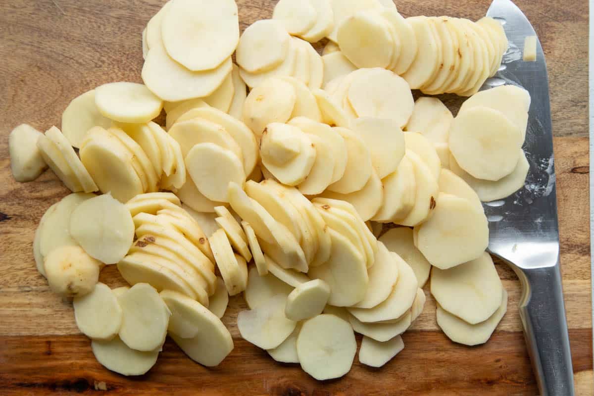 thinly sliced potatoes on a wooden cutting board next to a knife.