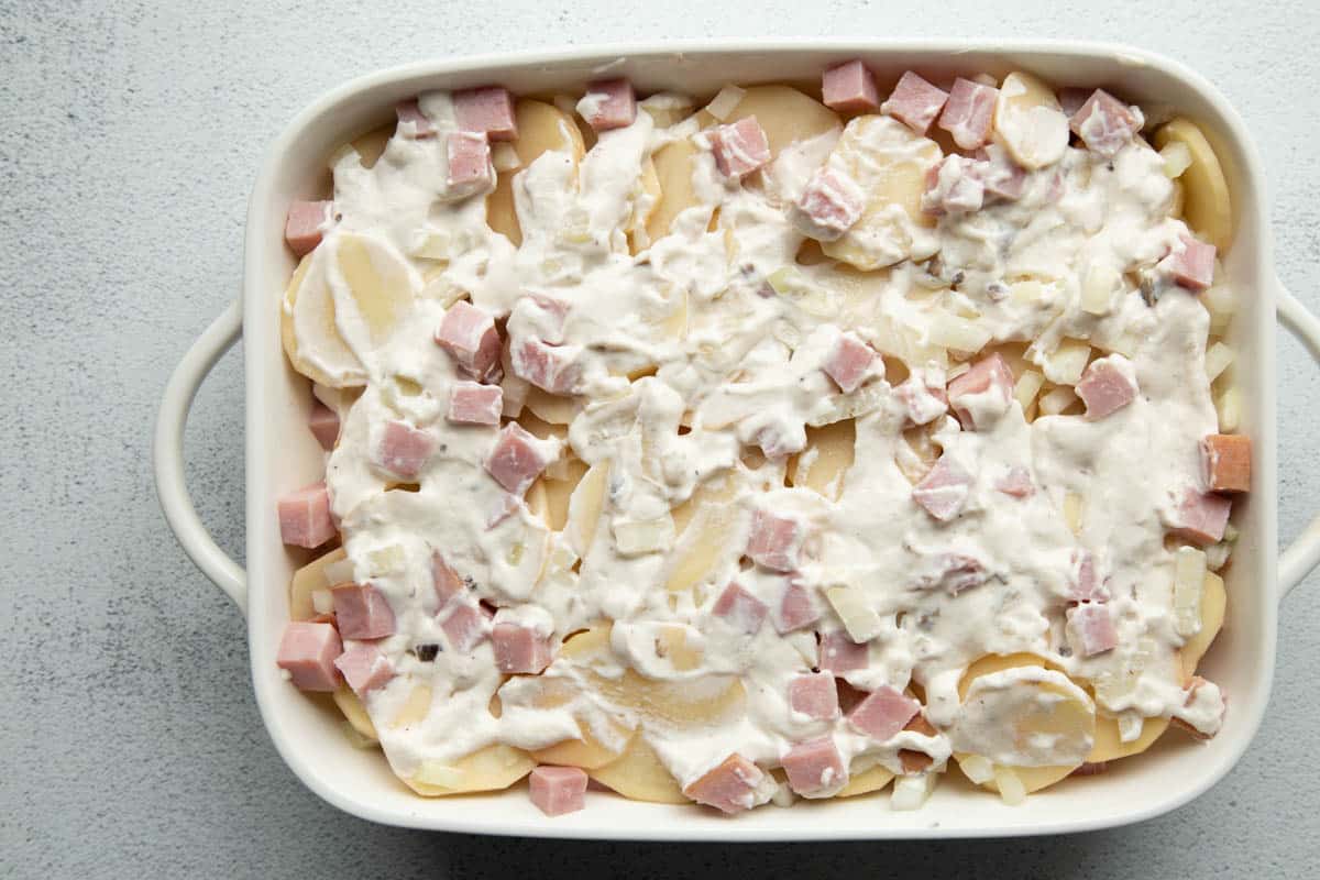 casserole dish with slices potatoes, diced ham, and a creamy sauce, shown unbaked.