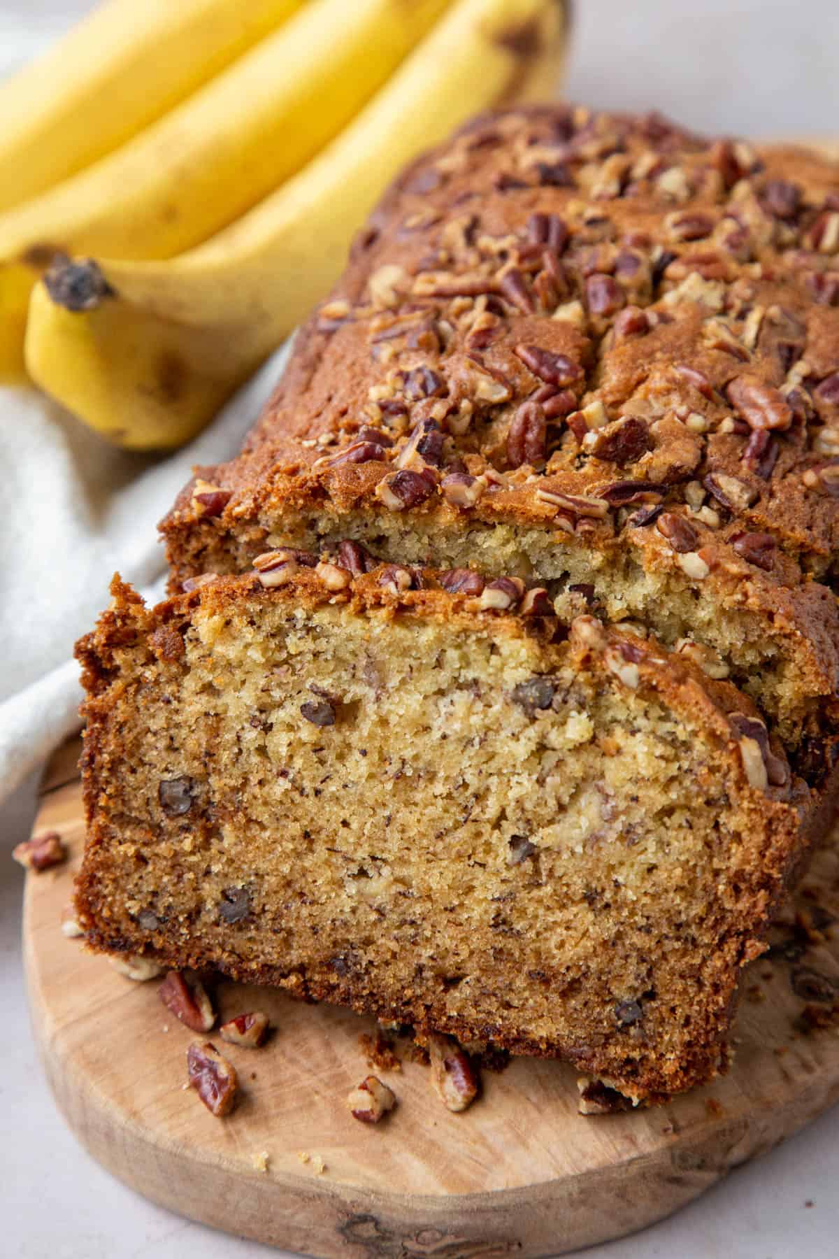banana bread loaf topped with pecans with slices cut out, next to a bunch of ripe bananas. 