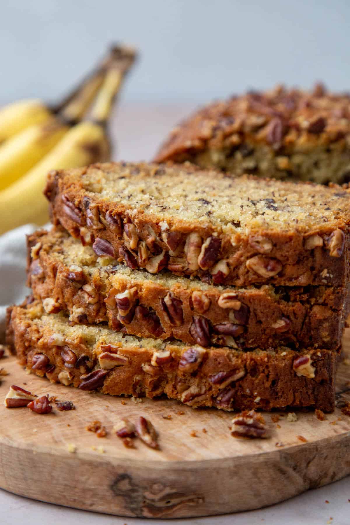 three slices of banana bread with pecans on top of a wooden serving board with ripe bananas in the background.