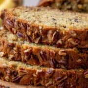 three slices of banana bread with pecans on top of a wooden serving board with ripe bananas in the background.