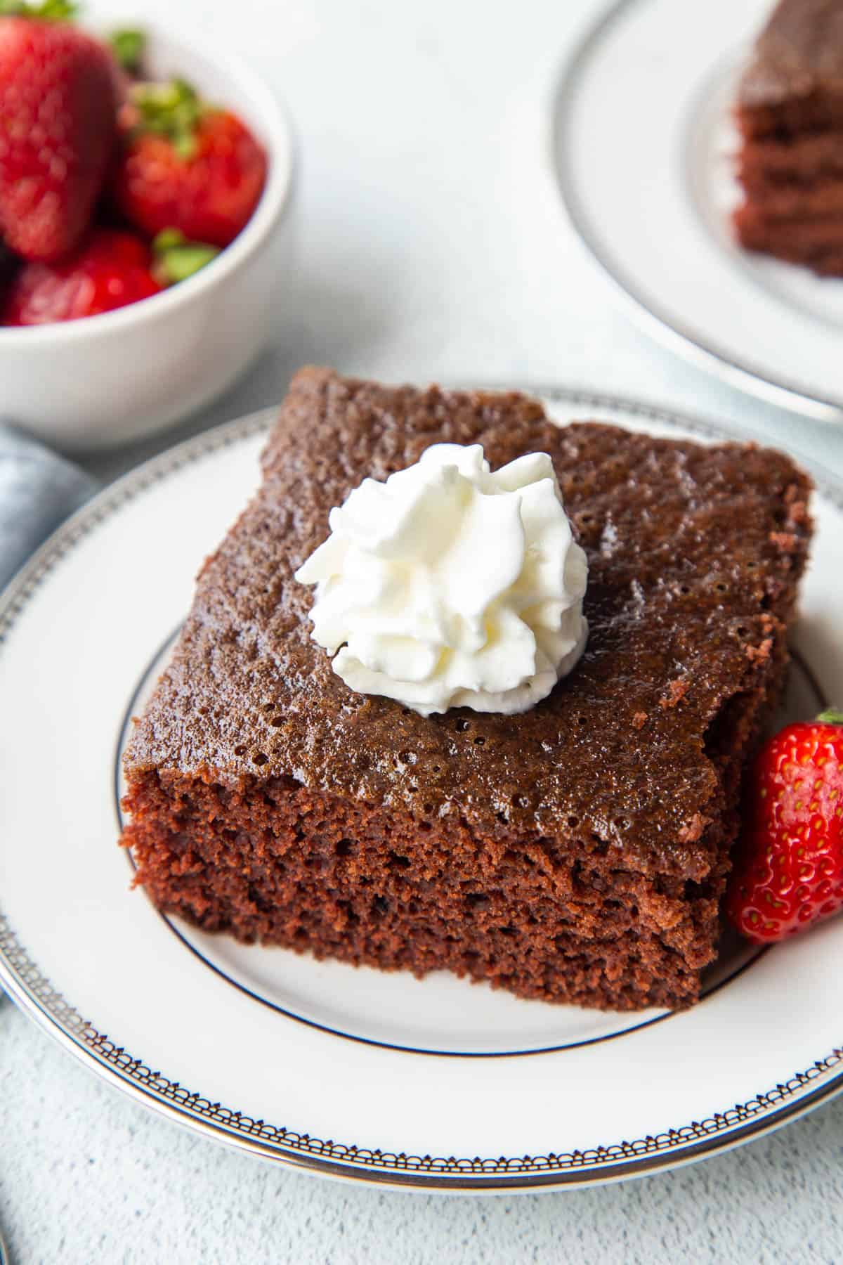 slice of wacky cake on a white plate, surrounded by strawberries and topped with whipped cream.