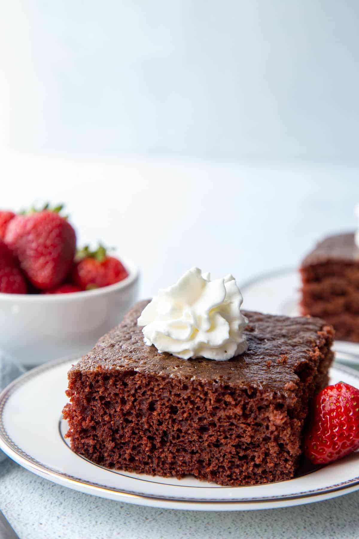 chocolate wacky cake slice topped with whipped cream on a white plate next to a few strawberries.