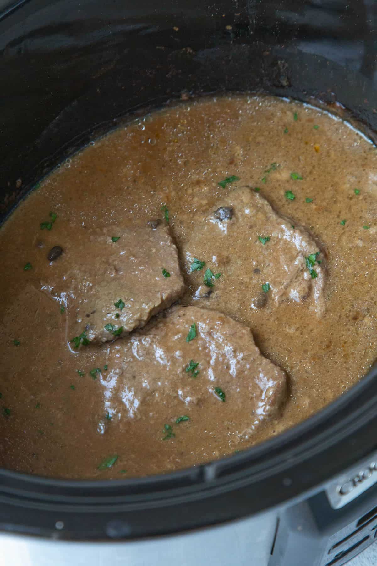 three cube steaks in a brown gravy in a slow cooker, topped with chopped parsley.